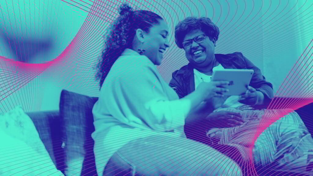 Two women smile at one another while using a tablet device